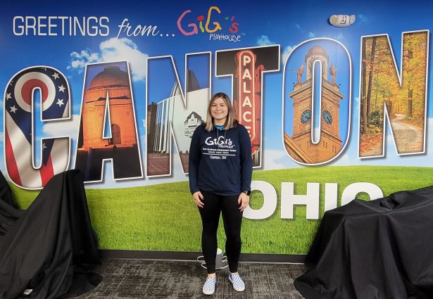 Photo of a GiGi's Playhouse volunteer wearing a GiGi's Playhouse Canton shirt in front of decorative Canton sign