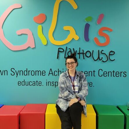 Claire, volunteer, sitting on colored cube chairs in front of GiGi's Playhouse sign
