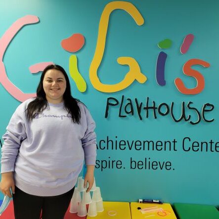 Girl in light purple sweater with long, dark hair in front of the GiGi's Playhouse logo