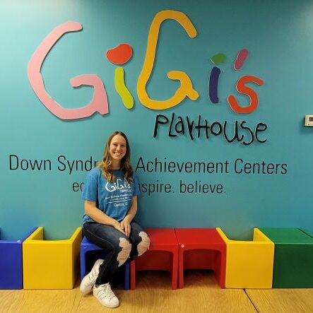 girl with blonde hair sitting in front of the GiGi's logo and wearing a blue GiGi's t-shirt