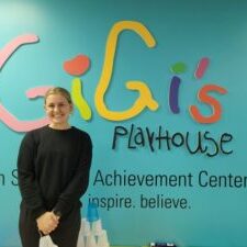 girl with blonde hair wearing all black in front of the colorful GiGi's Playhouse logo