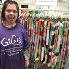 Girl with Down syndrome wearing a purple GiGi's Playhouse t-shirt in front of a rack of quilts.