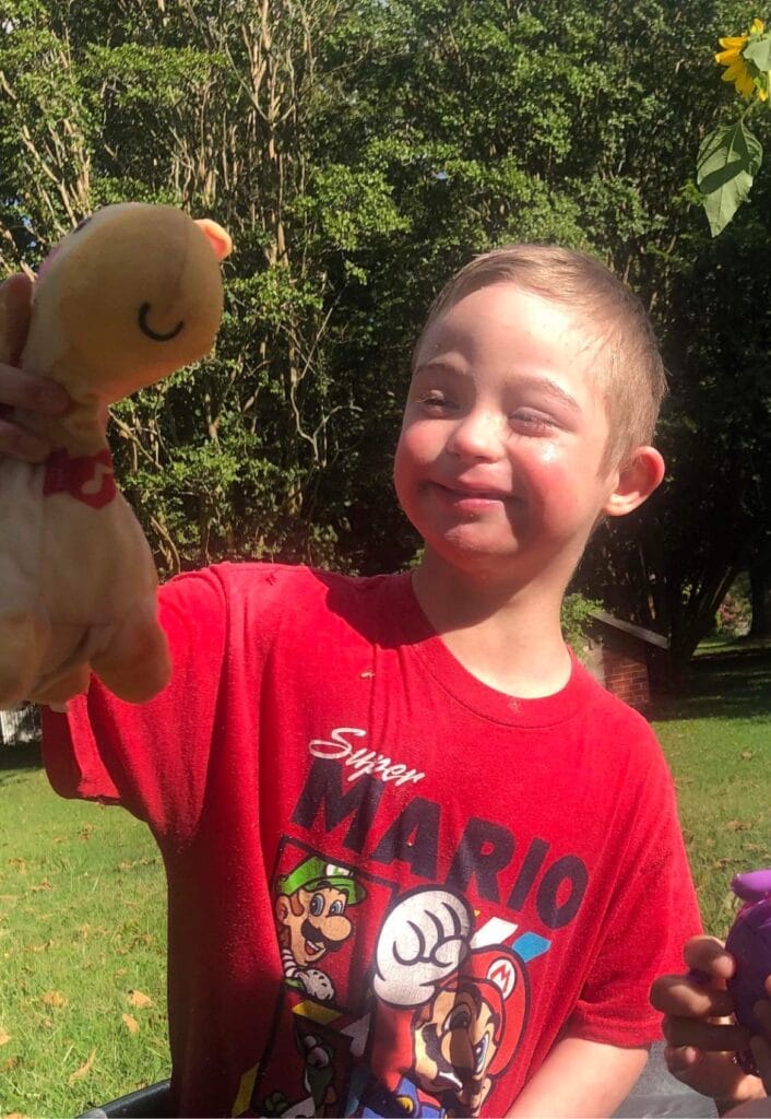 Young both with Down syndrome smiling at his stuffed animal while standing in the sunshine.
