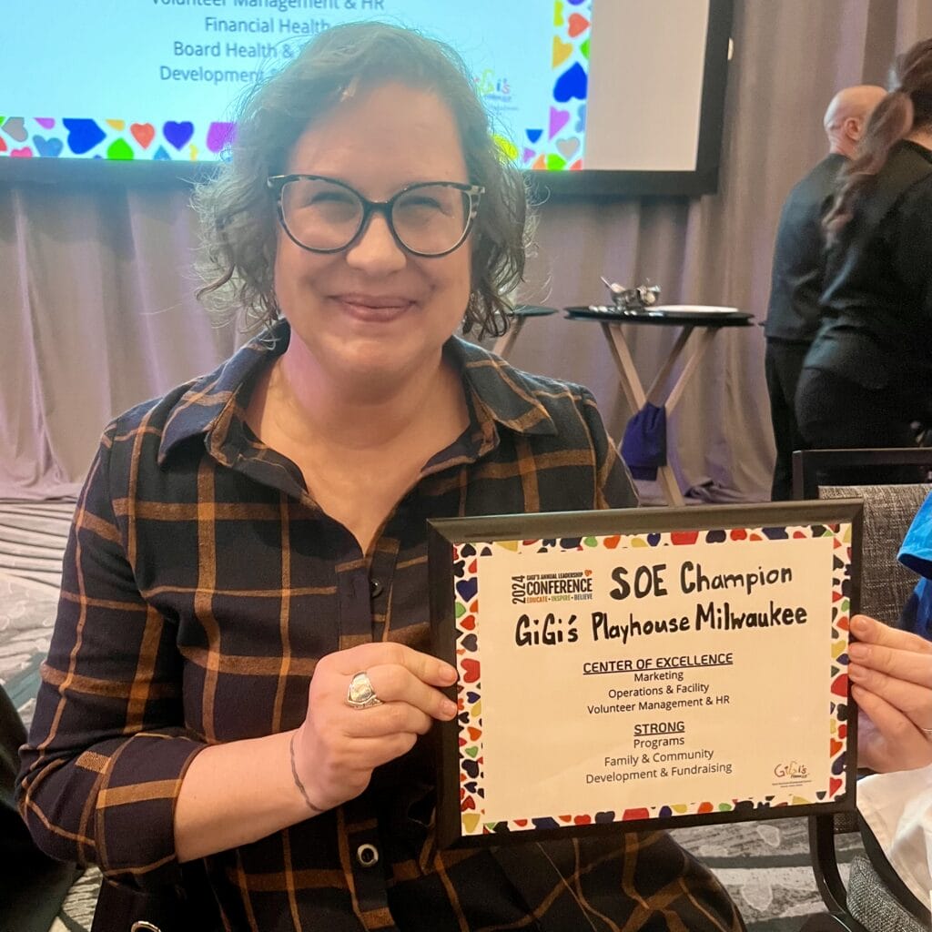 Woman holds a plaque noting Center of Excellence status for a job well done at GiGi's Playhouse Milwaukee.