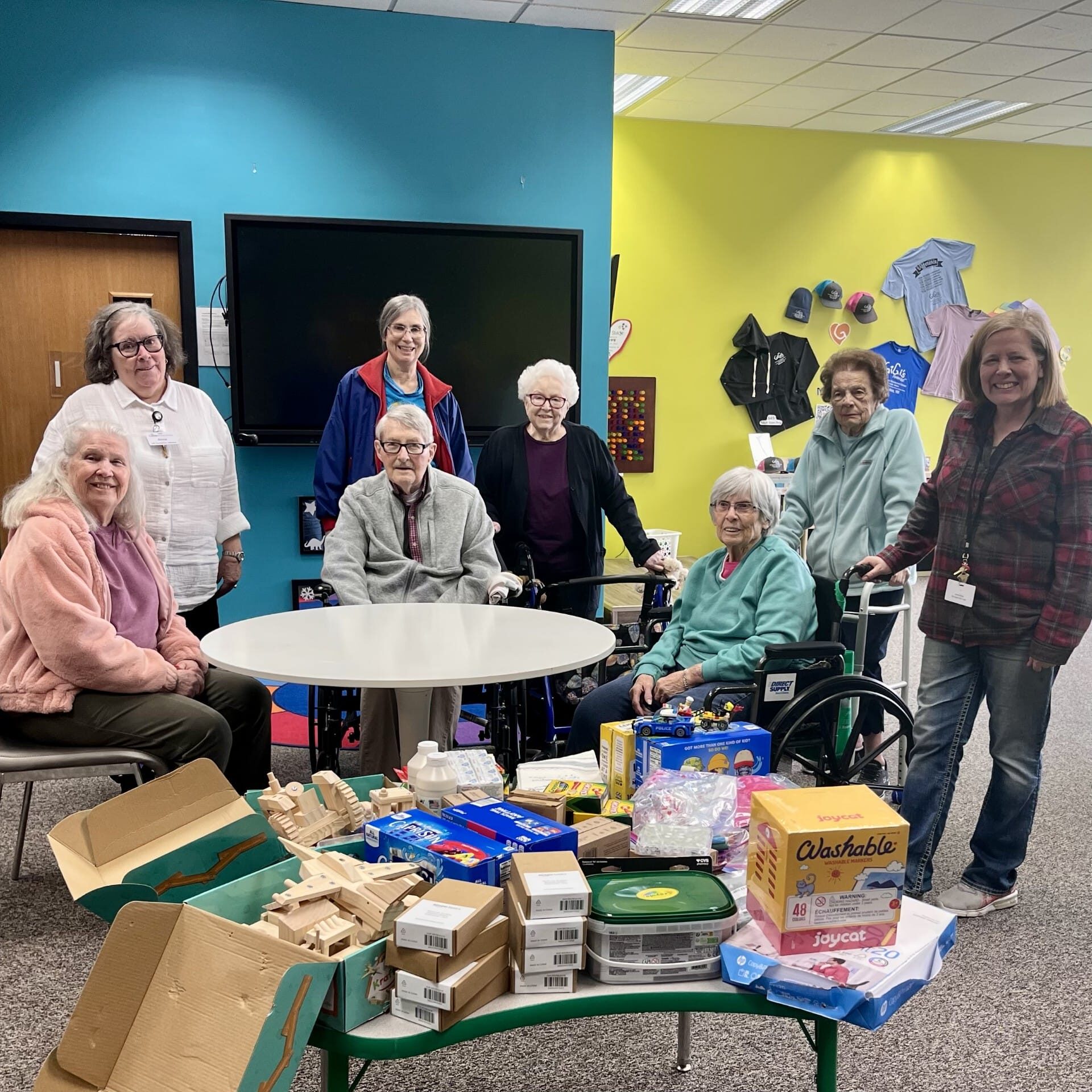 A group from Castle Senior Living poses at GiGi’s Playhouse Milwaukee, smiling around a table filled with donated art supplies, puzzles, and learning materials. The colorful Playhouse setting highlights this joyful moment of community giving.