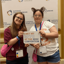 Mia and Mia pose for a photo at the National Down Syndrome Congress Conference. They are holding a sign that says "I met my BFF's at the NDSC Convention"