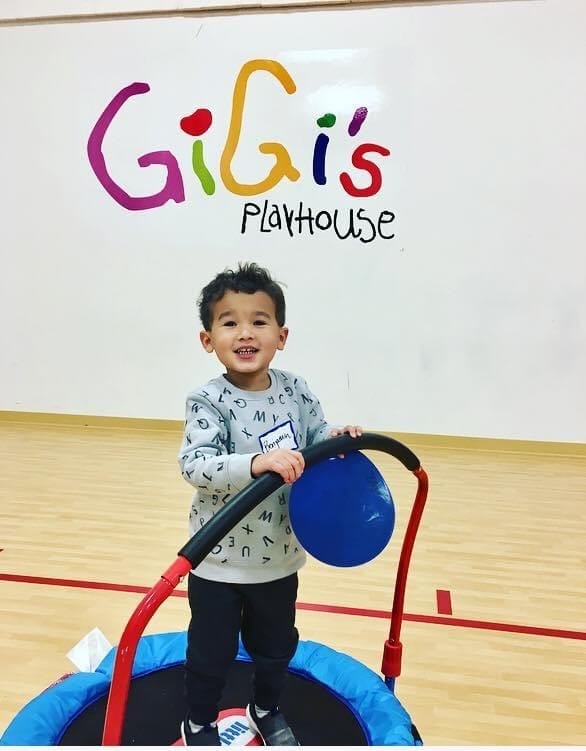 Young boy wearing  a gray sweater jumping on a small trampoline in a gym holding a blue dot in front of a GiGi's Playhouse Sign.