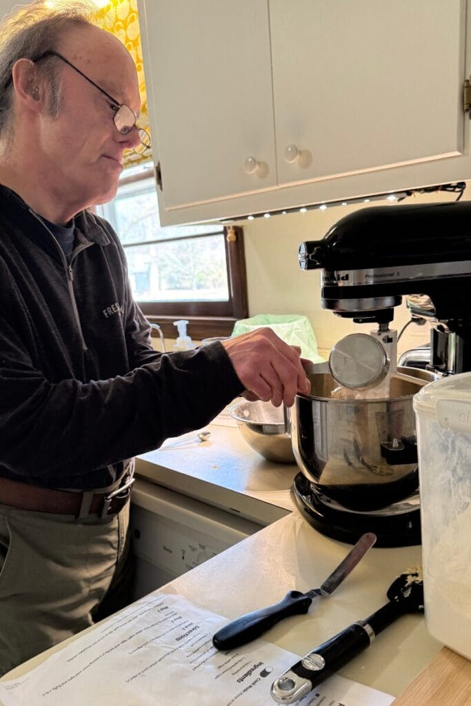 John pouring a cup of flour into a sliver mixing bowl.