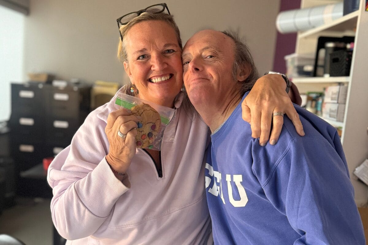 A women holding up an M&M cookie in a bag with her arm around an older gentleman with Down syndrome.