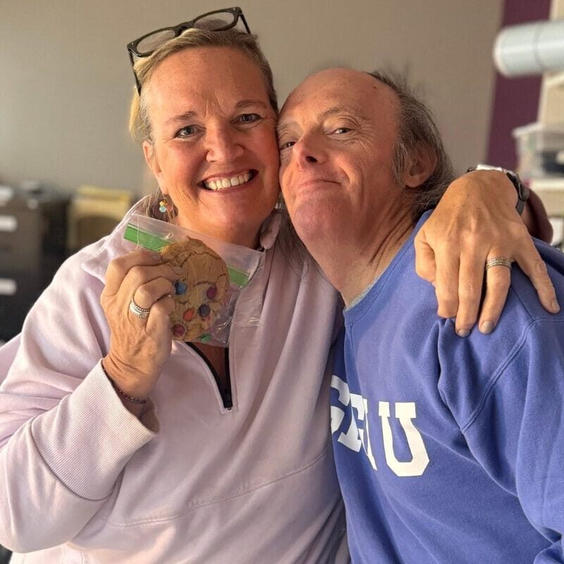 A women holding up an M&M cookie in a bag with her arm around an older gentleman with Down syndrome.
