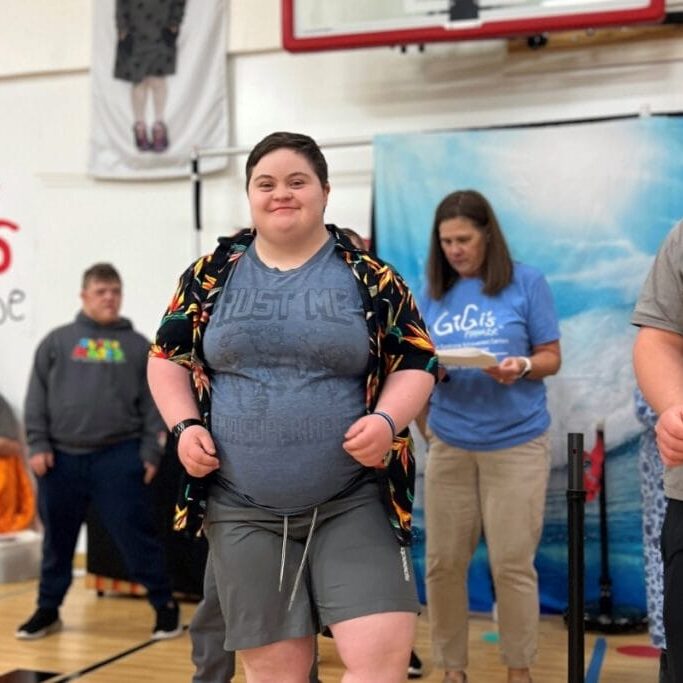 A girl with Down syndrome and short brown hair in a hawaiian shirt standing smiling at camera.