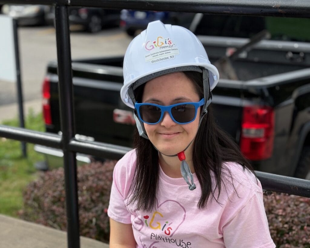 A smiling young adult with Down syndrome in a hard hat, symbolizing community and progress.